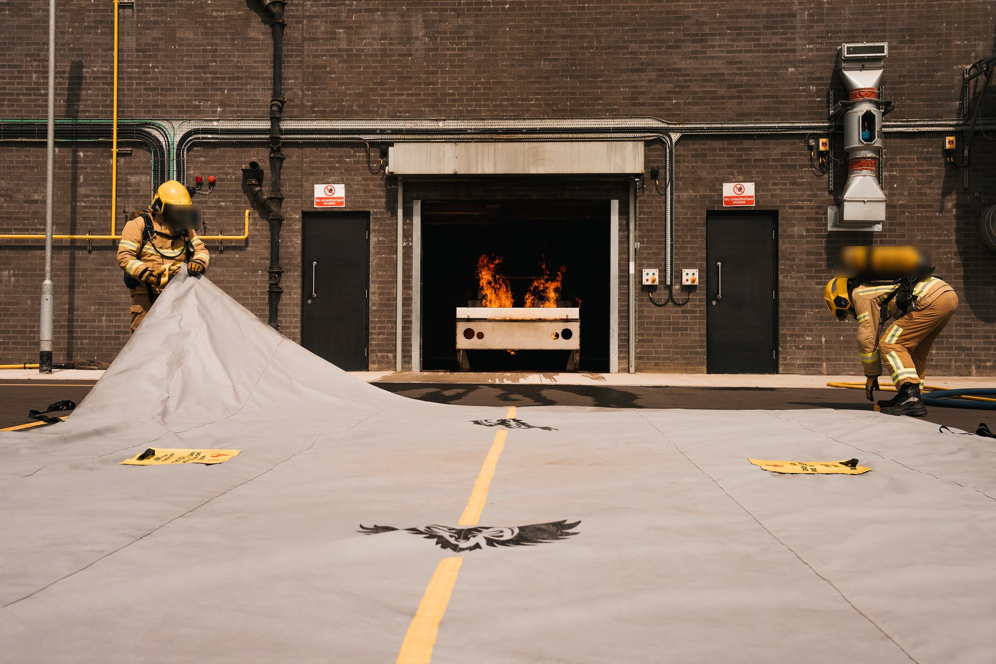 Two firefighters in full PPE preparing the Golden Fleece EV containment blanket in front of an active flame at a loading bay — real deployment of the EV thermal-runaway containment system, ram-and-wings brand mark and yellow alignment stripe visible on the spread blanket.
